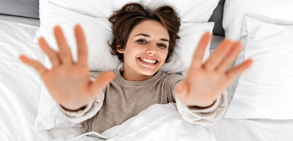Smiling woman lying on a white pillow and bedding, reaching both hands toward the camera, relaxed on a hypoallergenic mattress